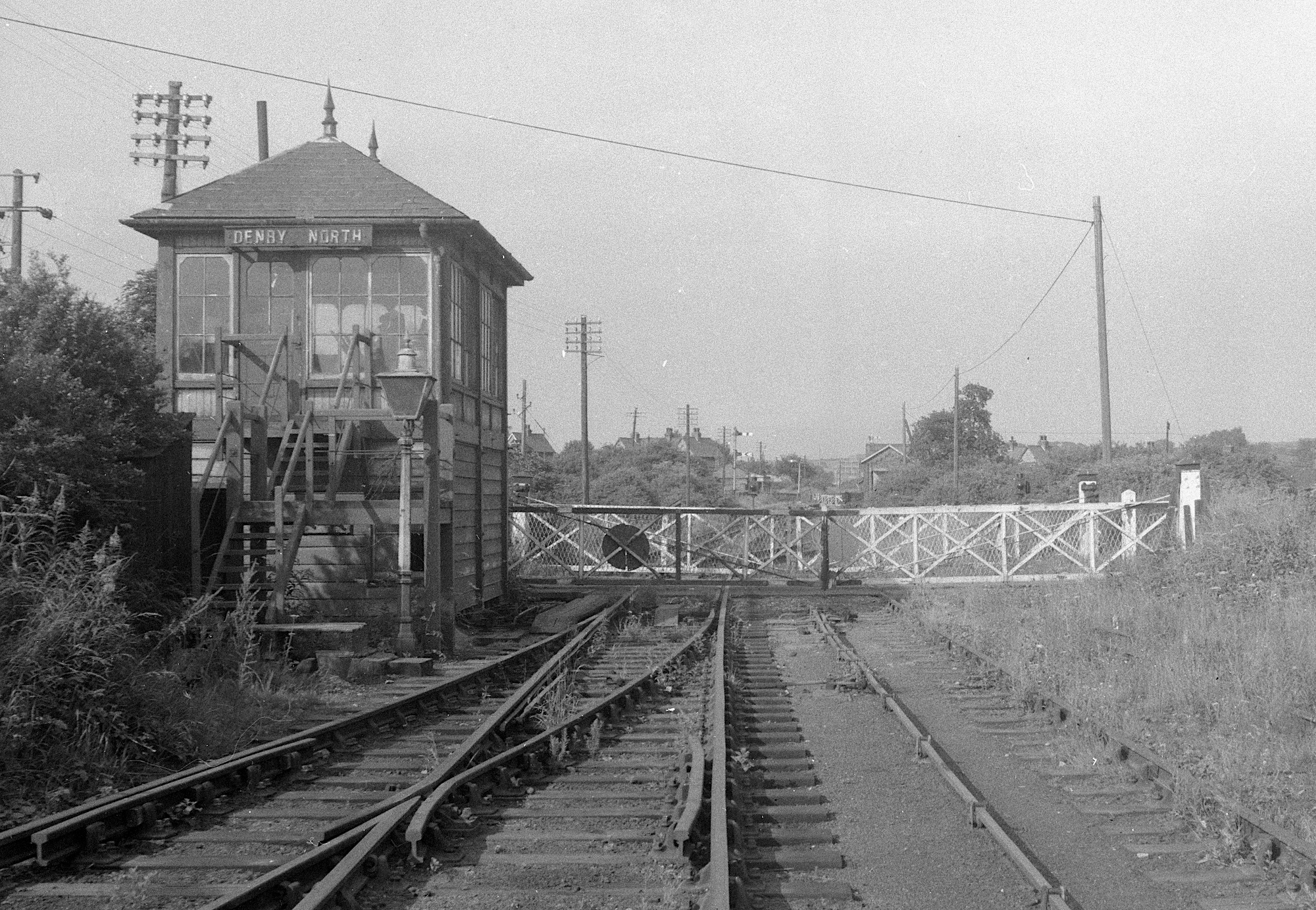 A black and white image taken from track level of a barely used railway line. Level crossing gates block the way and Denby North signal box fills the left side of the view