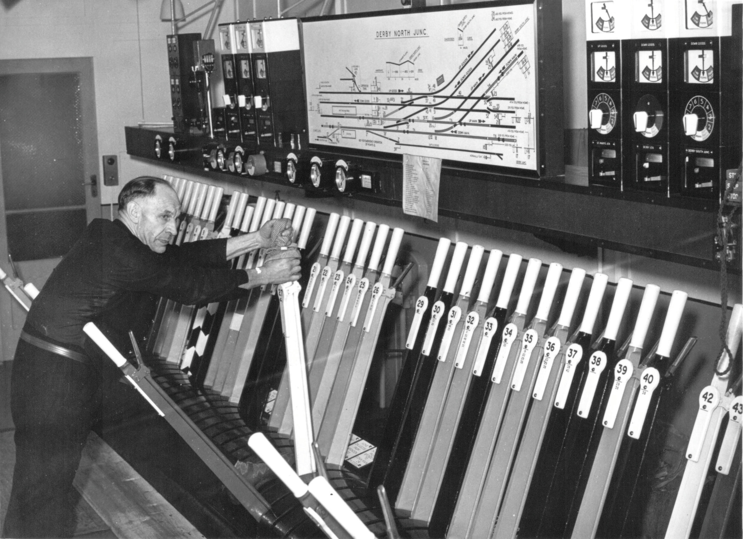 A Signalman in his 50s pulls one of over 40 levers visible inside the Derby North Junction signal box. Above the lever frame is a shelf filled with block instruments and an illuminated diagram of the track and signal layout.