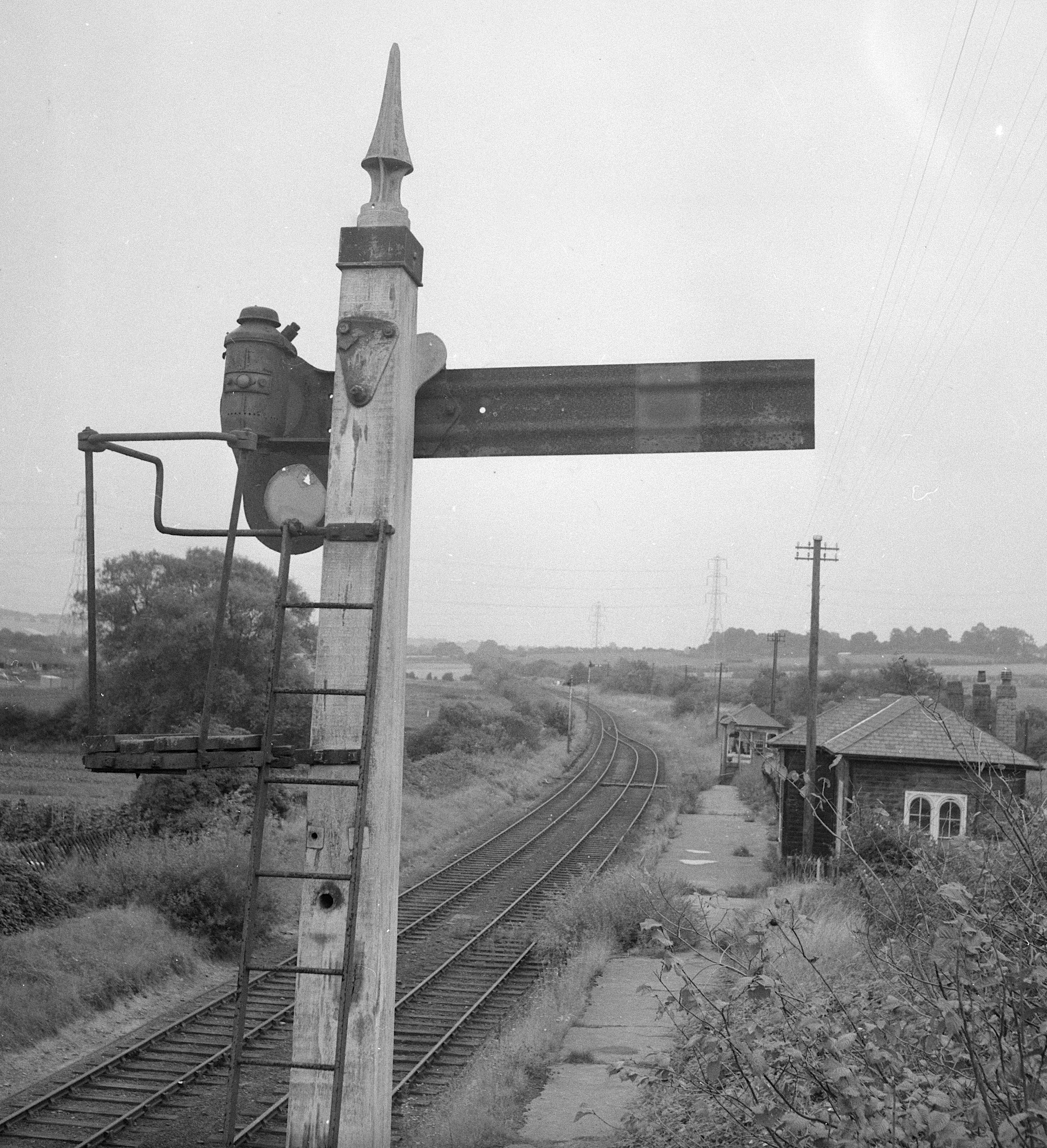A black and whte view from an overbridge showing the detail of the back of a Midland Railway Home Signal with it lamp arrangement. SOme near-disused startion buildings are in the background.